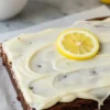 Homemade frosted lemon brownies with creamy lemon glaze on a white plate, garnished with lemon zest.